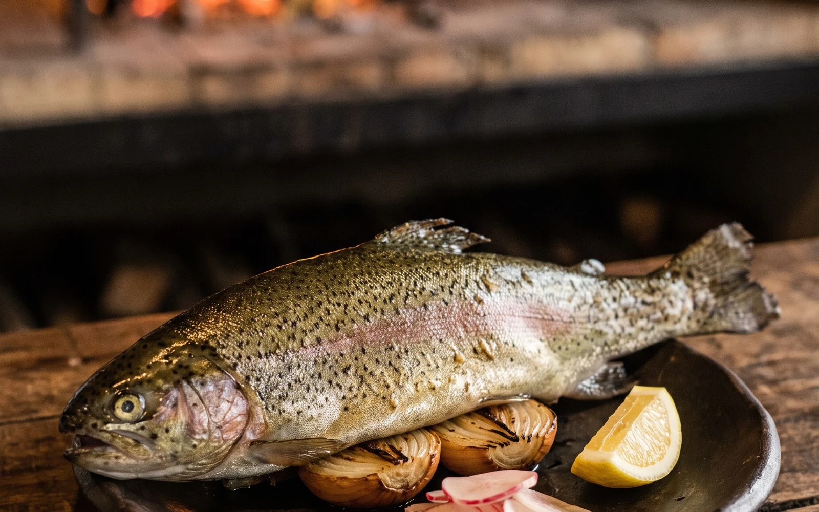 Whole ember-grilled wild trout with speckled skin, roasted onion and pickled radishes on a dark clay plate