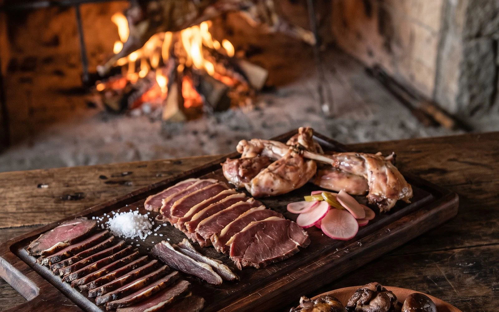 Wooden board with smoked slices, grilled lamb offal and a morel, with lamb over the fire behind