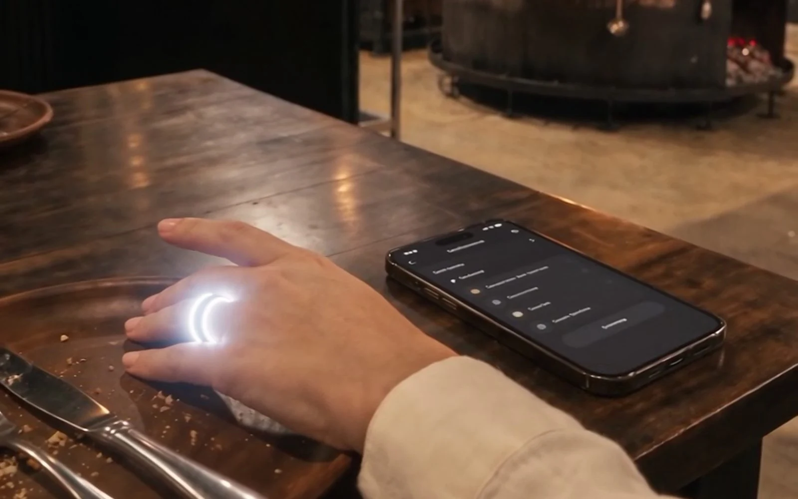 A hand wearing the lit biosensor ring on the table, beside the phone, with the hearth behind