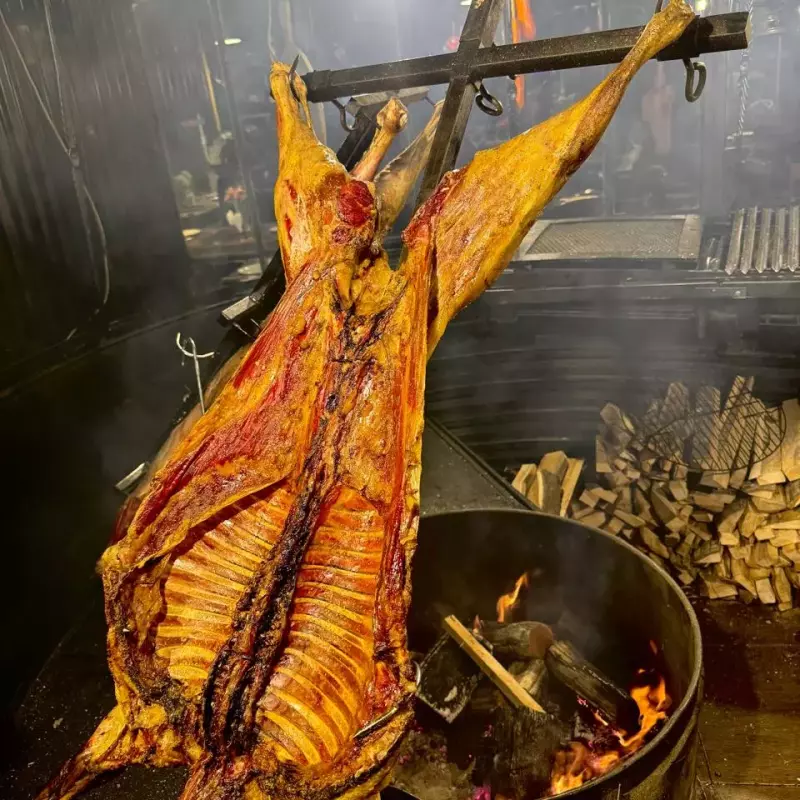 Real lamb opened on the cross over the embers, with stacked firewood beside it